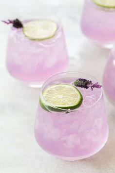 three glasses filled with lavender lemonade and limes on top of a white table