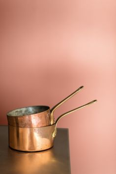 two brass pots sitting on top of a metal table next to a pink wall with gold spoons sticking out of it