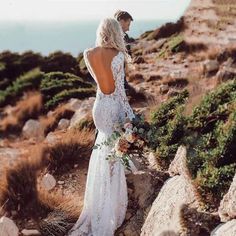 a woman standing on top of a rocky hillside wearing a wedding dress with open back