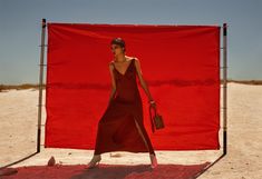 a woman in a long red dress is walking on the beach with a handbag