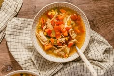 a bowl of soup with chicken and vegetables on a wooden table next to a spoon