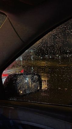 rain is falling on the windshield of a car at night with buildings in the background