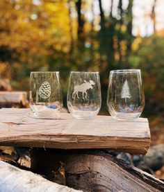 three wine glasses sitting on top of a piece of wood next to trees and leaves