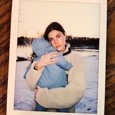 a woman holding a baby in her arms on top of a wooden table next to a snow covered field