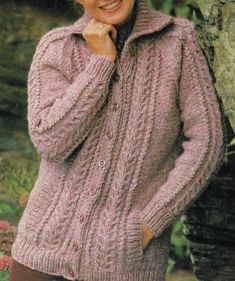 a woman wearing a pink sweater and brown pants standing in front of a tree with her hand on her chin