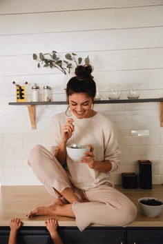 a woman sitting on top of a kitchen counter holding a cup and eating something out of a bowl