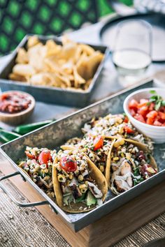 two tacos in a metal tray on a table next to bowls of salsa and chips