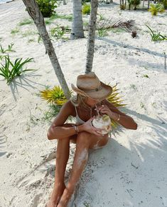 a woman sitting on the beach with a coconut in her hand and wearing a straw hat