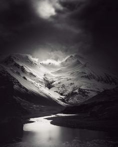 a black and white photo of a mountain range with water in the foreground under a cloudy sky