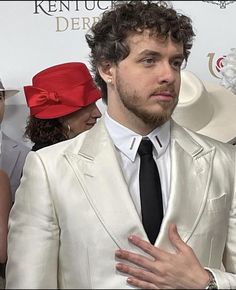 a man in a white suit and red hat with his hands on his chest looking at the camera