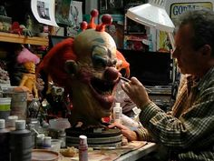 a man painting a creepy clown's face on a table in a room full of clutter