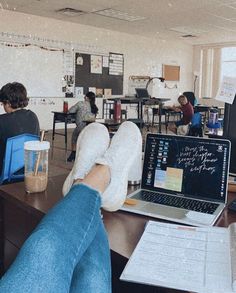 a person's feet resting on a desk in front of a laptop and other students