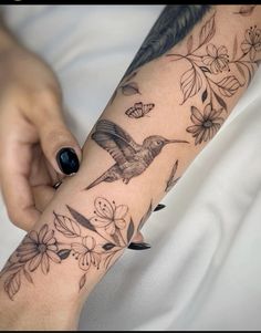 a woman's arm with tattoos on it and flowers around the wrist, in front of her is a hummingbird