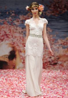 a woman walking down a runway wearing a white dress with flowers in it's hair