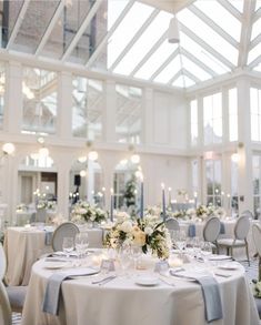 White round tables with hints of blue in the napkins and candles and a floral centerpiece in an atrium setting. Blue And White Flower Table Arrangements, Blue And White Round Table Decor, Wedding Settings Indoor, Wedding Table Decor Blue And White, Blue Round Table Wedding, Dusty Blue Wedding Venue Decor, Blue And White Table Setting Wedding, Blue And White Wedding Round Table Decor, Blue And White Wedding Reception Decor