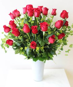 a vase filled with lots of red roses on top of a white table next to a plant