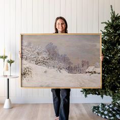 a woman holding up a large painting in front of a christmas tree with snow on it