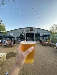 a person holding up a beer glass in front of a building with tables and umbrellas