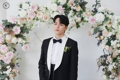 a young man in a tuxedo and bow tie standing next to a floral arch