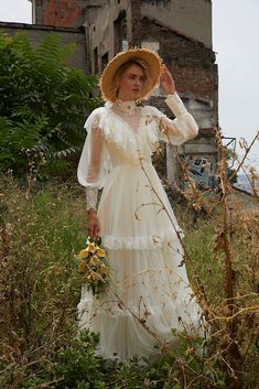 a woman in a white dress and straw hat is standing in tall grass near an old building