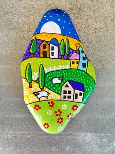 a painted rock sitting on top of a cement floor next to a wall with houses and trees