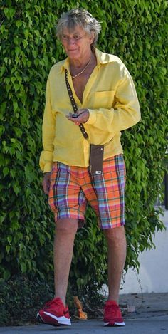 an older man in yellow shirt and plaid shorts standing next to a bush with cell phone