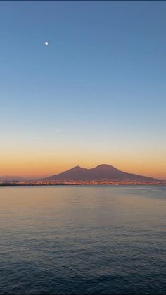 the sun is setting over an ocean with mountains in the distance and a crescented moon