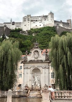 a man standing next to a fountain in front of a castle on top of a hill