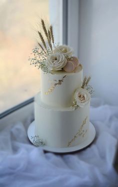 a three tiered white cake with flowers and feathers on the top is sitting in front of a window