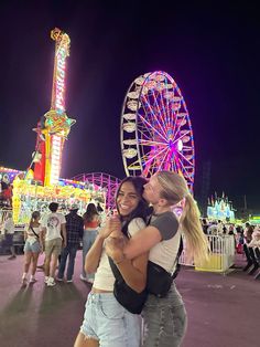 two women hugging each other in front of a ferris wheel at an amusement park during the night