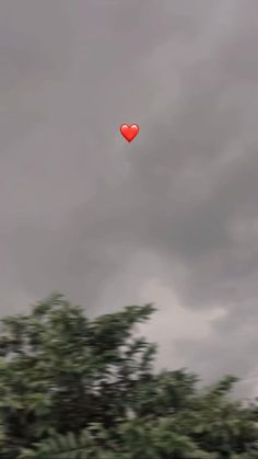 a red heart shaped balloon flying in the air over trees and clouds on a cloudy day