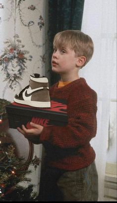 a young boy holding a shoe box in front of a christmas tree