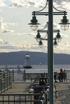 people are sitting on benches near the water with boats in the ocean and a lighthouse in the distance