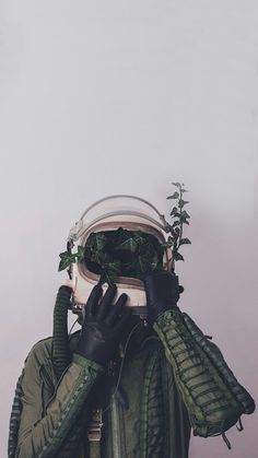 a person wearing black gloves and green jacket covering their face with plants growing out of it