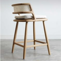 a wooden stool with a woven seat and backrest on concrete flooring, in front of a white wall