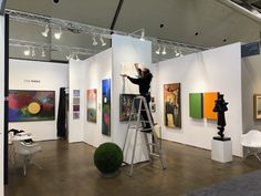 a man standing on a ladder in an art gallery painting pictures with his hands up