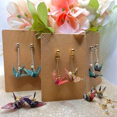 three different colored bird earrings on display in front of some pink flowers and green leaves