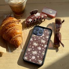 an iphone case sitting on top of a wooden table next to sunglasses and a croissant