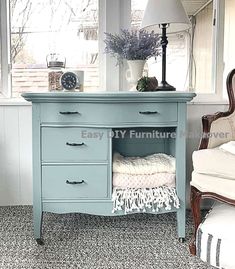 a blue dresser sitting in front of a window next to a white chair and lamp