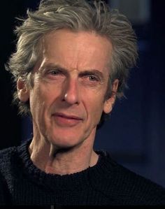 doctor who is looking at the camera while wearing a black sweater and grey hair, in front of a dark background