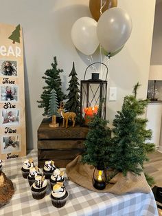 a table topped with cupcakes covered in frosting next to trees and balloons