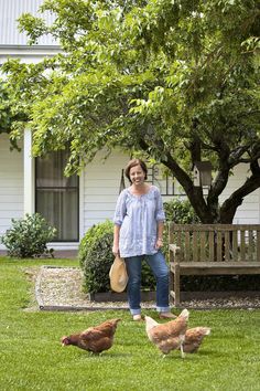 a woman standing in the grass next to two chickens and a bench with a tree behind her