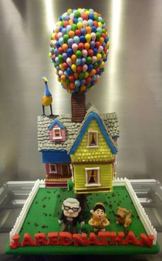 a cake that is shaped like a house with balloons in the air and people on it