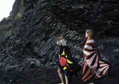 two women in colorful clothing walking on the beach next to a rocky cliff side area