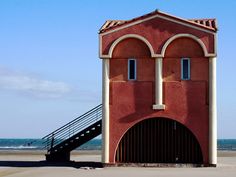 a tall red building sitting on top of a sandy beach next to the ocean with stairs leading up to it