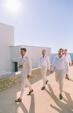 three men in white suits are walking down the sidewalk by the ocean with their backs to the camera