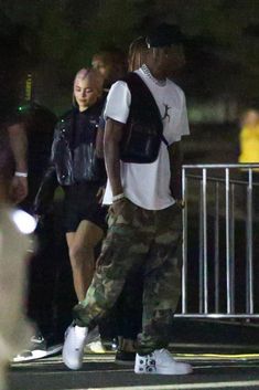 two people walking down the street at night, one is wearing a backpack and the other has a white t - shirt