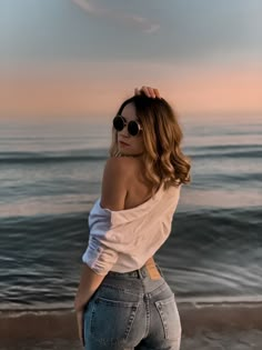 a woman standing on top of a beach next to the ocean wearing high waist jeans