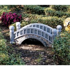 a small stone bridge in the middle of some bushes and flowers with purple flowers behind it