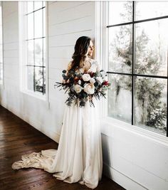 a woman standing in front of a window with flowers on her wedding dress and holding a bouquet